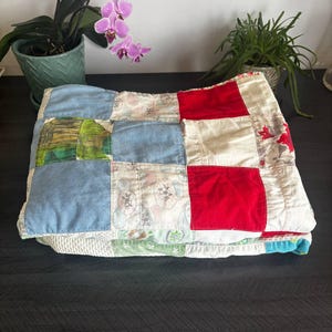 May include: Folded patchwork quilt with squares of blue denim, red, white, and patterned fabrics. The quilt is handmade and appears to be vintage. The quilt is folded neatly on a dark surface.