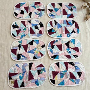 May include: A set of eight oval-shaped fabric coasters featuring a patchwork design of various geometric shapes in shades of pink, blue, white, black, and maroon, with visible white stitching outlining each individual fabric piece and the edges.
