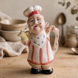 May include: A ceramic jug shaped like a chef wearing a white hat with red trim, a white apron with blue dots and red vertical stripes, and black shoes. The chef figure holds a small yellow jug in one hand and has a handle on the back.