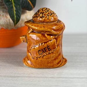 Op de afbeelding: Een bruine keramische bus in de vorm van een jute zak met een textuurrijk oppervlak. Het deksel is gevormd als een hoop geroosterde koffiebonen. Een rechthoekig label aan de voorzijde bevat de zwarte tekst CAFÉ in een schreefloos lettertype.