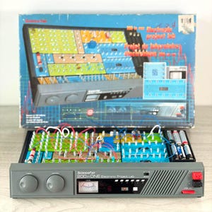 Puede incluir: Un kit Science Fair 200 in ONE Electronic Project Lab que presenta una consola gris con dos perillas grandes, un medidor de nivel y una tecla de código roja. El kit incluye una placa de circuito colorida con varios componentes y cables.