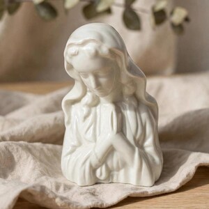 May include: A white ceramic bust of a figure with a draped head covering, depicted with eyes closed and hands pressed together in a prayer position. The sculpture features smooth, glossy surfaces and detailed folds in the fabric-like garment.