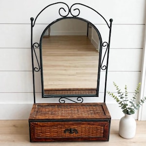 Peut inclure: Un miroir de courtoisie mural doté d'un cadre en fer forgé noir avec un sommet arqué et des volutes décoratives. Sous le miroir se trouve une boîte de rangement rectangulaire en bois avec une façade en osier tressé et une poignée en métal.