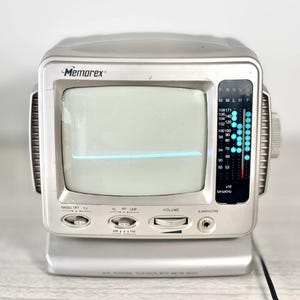 May include: A silver Memorex mini personal television with AM/FM radio. The screen shows a horizontal blue line. Features include volume control, earphone jack, and radio frequency dial. The text "Memorex" is visible on the top.