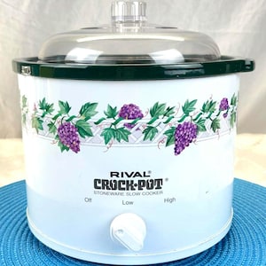 May include: White Rival Crock-Pot stoneware slow cooker with a clear glass lid and a dark green rim. The slow cooker features a decorative band of purple grapes and green leaves. The control knob has settings for off, low, and high.