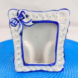 Peut inclure: Un petit cadre photo en céramique avec un fond blanc et une bordure bleue. Le cadre a un nœud en haut et est décoré de pois verts. Le cadre mesure environ 10 cm de haut et 7,5 cm de large.