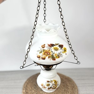 May include: A vintage hanging lamp with a white glass shade and base, decorated with floral patterns in yellow, brown, and white. The lamp is suspended by a dark metal chain.