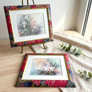 Ett par blomlitografier av Laura Ash, irisar och orkidéer inramade i vintageramar täckta med blommönstrat tyg.