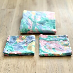 Peut inclure: Trois pièces de literie avec un motif floral à l'aquarelle dans les tons de bleu, rose, vert et jaune. Le tissu est plié et disposé sur une surface en bois brun clair. La literie semble être un ensemble de draps.