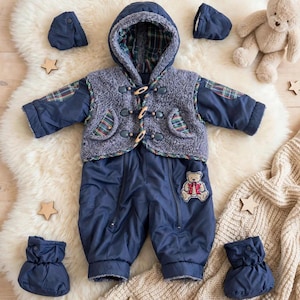 Op de afbeelding: Een marineblauw sneeuwpak met capuchon, voorzien van een grijs sherpa fleece vest met houtje-touwtjesluiting, ruitpatroon accenten op de mouwen en zakken, en een kleine geborduurde teddybeer-patch op het been, inclusief bijpassende wanten en slofjes.