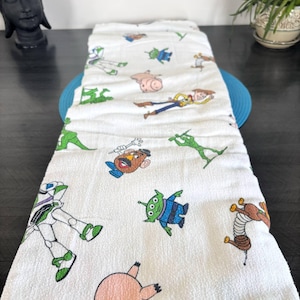 Może przedstawiać: Biały bawełniany ręcznik z kolorowymi postaciami z kreskówki z filmu Toy Story. Ręcznik ma wizerunki Buzza Astrala, Chudego i innych postaci. Ręcznik jest złożony i umieszczony na niebieskiej macie.