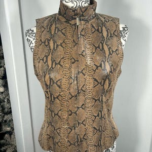 May include: A sleeveless vest with a snakeskin pattern in shades of brown and black. The vest has a high collar and a silver zip down the front. The garment is displayed on a mannequin.