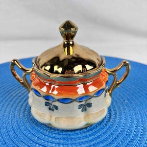Peut inclure: Un sucrier orné avec un couvercle et des poignées dorés. Le bol présente une base blanche avec des motifs floraux bleus, une bande orange et bleue et un bord doré. Le couvercle est surmonté d'un bouton doré.