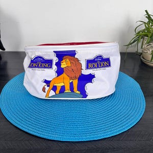 Peut inclure: Trousse de maquillage blanche avec un lion de dessin animé du Roi Lion. Le sac a une fermeture éclair rouge et les mots "The Lion King" et "Roi Lion" imprimés dessus. Le sac est posé sur un dessous-de-plat bleu.