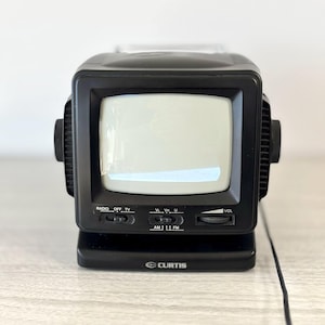 Puede incluir: Un televisor portátil Curtis negro vintage con una pantalla pequeña. El televisor tiene una función de radio y control de volumen. El texto "CURTIS" está impreso en la parte delantera.
