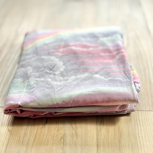 Pode incluir: Cobertor dobrado com um design pastel de arco-íris e nuvens. As cores incluem rosa, cinza e verde claro. O cobertor está cuidadosamente dobrado, mostrando o padrão completo. O fundo é uma superfície de madeira clara.