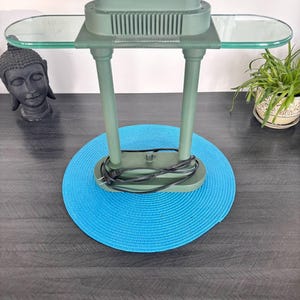 Peut inclure: Une lampe de bureau verte avec une étagère en verre et un dessus rectangulaire. La lampe a deux pieds de support et une base ronde. La lampe est posée sur un dessous-de-plat bleu. Un cordon noir est attaché à la lampe.