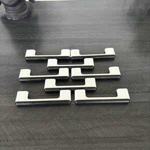 Peut inclure: Six poignées de meubles au design moderne. Chaque poignée présente une section centrale rectangulaire blanche avec des extrémités noires incurvées. Les poignées sont disposées sur une surface gris foncé, suggérant un thème de mobilier ou de quincaillerie.
