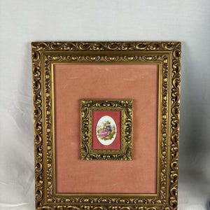 Puede incluir: Un cuadro en miniatura enmarcado que representa una escena romántica. La pintura está dentro de un marco dorado más pequeño y ornamentado, que está centrado en un marco dorado más grande con un passe-partout de color coral. Los marcos tienen tallas decorativas detalladas.
