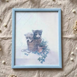 Peut inclure: Une impression encadrée représentant deux chatons, un gris clair et un tabby foncé, assis dans un seau en bois marron. Un petit pot en terre cuite avec des feuilles de lierre vert se trouve à côté du seau, avec des tiges tombant devant.
