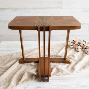 May include: A rectangular wooden side table with a smooth brown finish and a central decorative inlay featuring a dark, intricate pattern. The table is supported by a unique base design consisting of three vertical slats connected to two legs.