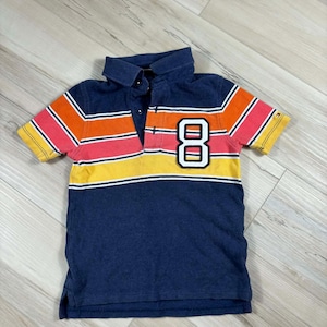 Peut inclure: Polo bleu marine avec un motif color block à rayures orange, rouge et jaune sur la poitrine et les manches. Le chiffre "8" est affiché en blanc sur la poitrine. Le polo a un col classique et des manches courtes.