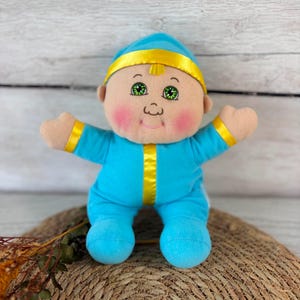Peut inclure: Une poupée en peluche avec une tête ronde, des yeux verts et des joues roses, portant une grenouillère bleu vif avec un bandeau en satin jaune et une bande jaune verticale au centre de la poitrine. La poupée a les bras et les jambes écartés.