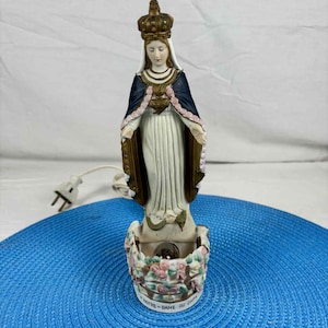 May include: A ceramic statue of the Virgin Mary, with a crown and a blue cloak with pink trim. The statue is mounted on a base decorated with flowers and the text "NOTRE-DAME DU CAP". A power cord and plug are visible.