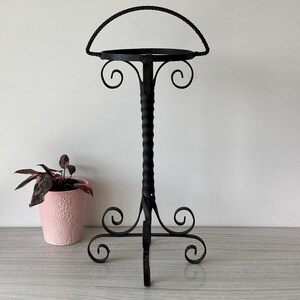 May include: Black wrought iron plant stand with a twisted central column and decorative scrollwork. The stand has a circular top and a curved handle. A pink potted plant is to the left of the stand.