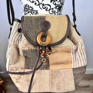 Peut inclure: Sac à dos patchwork aux tons neutres. Le sac présente une variété de textures, notamment du tissu tissé, du liège et de la toile. Il possède une fermeture à rabat avec un anneau en bois et une attache décorative en bois. Accents en cuir marron.