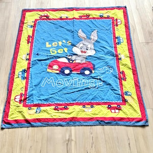 Op de afbeelding: Een blauwe, gele en rode babydeken met Bugs Bunny die in een rode auto rijdt. De deken heeft een rand met kleine auto-illustraties en de tekst "Let's Get Moving!"