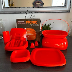 Peut inclure: Un ensemble de pique-nique vintage en plastique rouge, comprenant des assiettes, des tasses, des ustensiles et un récipient avec une poignée. La boîte indique "Picnic Master". L'ensemble est disposé sur une surface sombre.