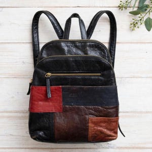 May include: A black leather backpack featuring a patchwork design with panels in red, dark brown, and tan. The bag includes a top handle, two shoulder straps, and a horizontal zippered front pocket with a gold-toned metal zipper pull.
