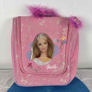May include: A pink Barbie travel bag featuring a Barbie image. The bag has a floral design and is adorned with pink feather trim. It has a zip closure and a handle. The Barbie logo is displayed on the front.