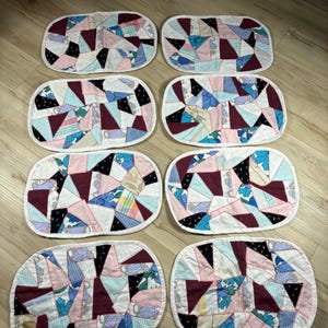 Peut inclure: Huit sets de table matelassés de forme ovale. Chaque set de table présente un motif patchwork avec des tissus bordeaux, bleus, roses et blancs. Les bords sont finis avec une bordure blanche. Les sets de table sont disposés sur une surface en bois.