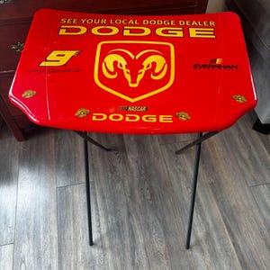 Peut inclure: Table pliante rouge avec le logo Dodge et du texte. La table présente un emblème de tête de bélier jaune et les mots "SEE YOUR LOCAL DODGE DEALER". La table a des pieds en métal noir.