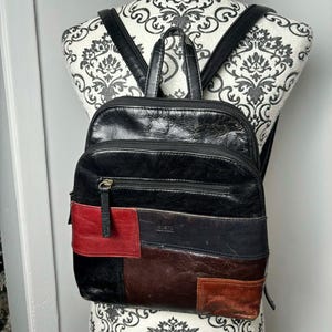 May include: Black leather backpack with a patchwork design featuring red, navy, and brown panels. The backpack has a top handle, adjustable straps, and multiple zippered compartments for storage. The bag is displayed on a decorative mannequin.