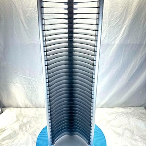 Può includere: Una torre per CD color argento con un design curvo, che contiene più CD. La torre è su un tappetino circolare blu. Il marchio "ATLANTIC" è visibile sulla base. La torre è realizzata in metallo e plastica.