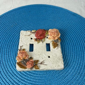 Puede incluir: Una cubierta doble para interruptor de luz de color blanco roto con una superficie texturizada. La cubierta está adornada con rosas esculpidas en tonos melocotón y rojo, junto con hojas verdes. La placa del interruptor se encuentra sobre un fondo azul texturizado.