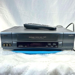 May include: A gray VCR player with a remote control on top. The VCR has a front panel with buttons and a display. The text "DA4 HEAD UNIVERSAL REMOTE HI-FI" is printed on the front.