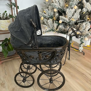 Op de afbeelding: Een antieke, donkergrijze kinderwagen met bijpassende kap. De wagen heeft een metalen frame met decoratieve krullen en spaakwielen. De kinderwagen is een decoratief item.