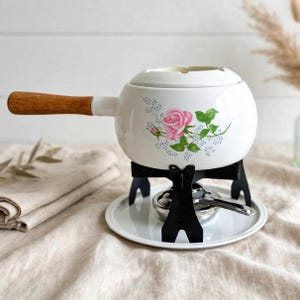Puede incluir: Una olla de fondue de cerámica blanca con mango de madera y un patrón floral de rosas rosas. La olla descansa sobre un soporte de metal negro con base circular y un pequeño quemador de metal debajo, diseñado para calentar el contenido.