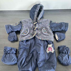 Peut inclure: Combinaison de neige pour bébé bleu marine avec veste à capuche et moufles et bottillons assortis. La veste a un gilet en polaire grise avec des fermetures à bascule en bois et des accents écossais. Un écusson ours en peluche est sur le devant.