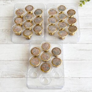 Peut inclure: Une collection de boutons de placard ronds dotés d'une base en métal doré et d'une incrustation circulaire au motif de pierre marbrée dans les tons marron et gris. Les boutons sont organisés et présentés dans trois plateaux de rangement en plastique transparent.