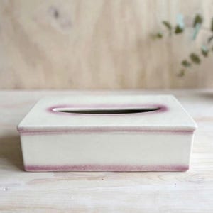 Op de afbeelding: Een rechthoekige keramische tissueboxhouder met een glanzende off-white afwerking. De randen van de opening aan de bovenzijde en de onderkant van de box zijn voorzien van een zachte, paars-roze kleurverloopglazuur voor een subtiele rand.
