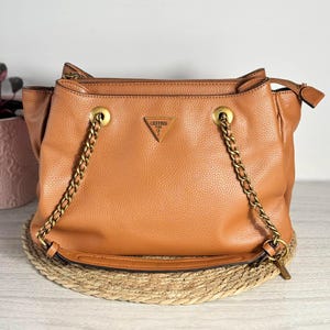 May include: A tan Guess handbag with gold-tone chain straps and hardware. The bag features a textured exterior, a top zipper closure, and a small Guess logo on the front. The bag is sitting on a woven mat.