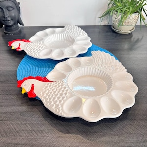 May include: Two white ceramic egg serving platters shaped like chickens. Each platter has a central bowl and egg-shaped indentations. The chicken heads are red and yellow, and the platters are on a blue mat.