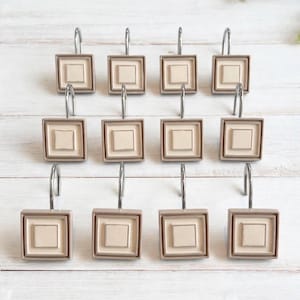 Peut inclure: Un ensemble de douze crochets de rideau de douche carrés présentant un carré central beige clair encadré par une bordure beige légèrement plus foncée, chacun fixé à un crochet en métal argenté poli pour une tringle de douche standard.