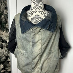 May include: A denim jacket with black leather sleeves and a large, dark blue collar. The jacket has a faded, worn appearance and features two front pockets. The jacket is displayed on a mannequin.
