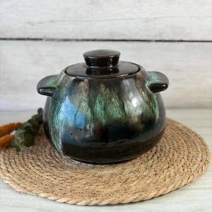 May include: A round ceramic lidded pot featuring a glossy dark brown base glaze with vibrant turquoise and teal drip patterns cascading down the sides. The vessel includes two side handles and a matching circular lid with a central knob handle.